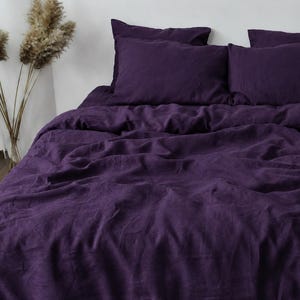 Könnte beinhalten: Ein tiefviolettes Bettwäsche-Set bestehend aus einem Bettbezug und zwei passenden Kissenbezügen. Der Stoff weist eine sichtbare, strukturierte Webart und ein leicht zerknittertes Aussehen auf. Die Bettwäsche liegt ordentlich auf einem Bettgestell.