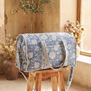 May include: A blue and white floral patterned duffel bag with a shoulder strap sits on a wooden stool. The bag has a cylindrical shape and is made of a quilted fabric. A straw hat and leather journal are nearby.