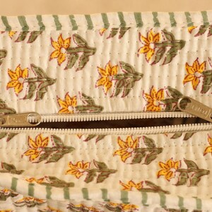 May include: Close-up of a cream-colored fabric with a floral pattern in yellow and green. The fabric has a quilted texture and a zipper closure. The zipper is silver-colored.