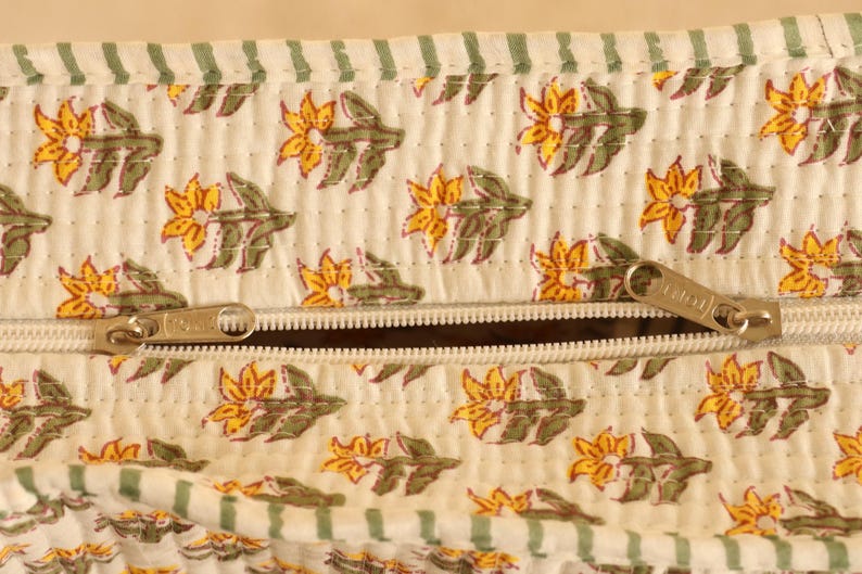 May include: Close-up of a cream-colored fabric with a floral pattern in yellow and green. The fabric has a quilted texture and a zipper closure. The zipper is silver-colored.