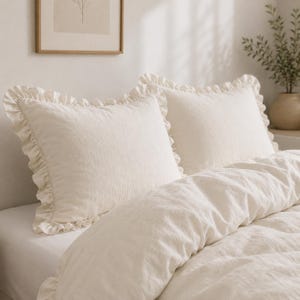 Può includere: Due federe rettangolari color bianco sporco con bordo arricciato. Il tessuto presenta una trama visibile e strutturata. I cuscini sono posizionati su un letto con un copripiumino abbinato, creando un set da letto coordinato.
