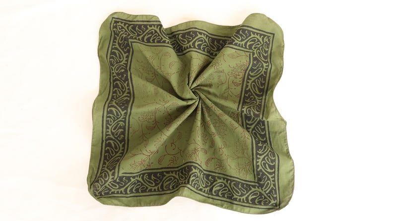 May include: A square olive green bandana featuring a dark brown border with a repeating paisley pattern and a central field decorated with a subtle, thin-lined brown floral motif. The fabric is folded in the centre, creating a soft, gathered texture.