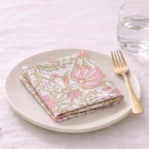 Può includere: Una tovagliolo bianco piegato con un motivo floreale rosa è appoggiato su un piatto bianco. Una forchetta dorata e un bicchiere d'acqua sono accanto al piatto. Il tovagliolo ha un design floreale rosa e verde.