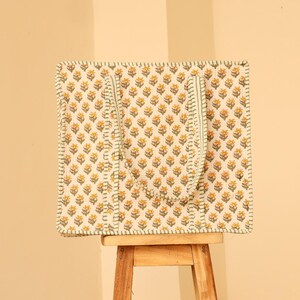 May include: A square tote bag with a floral pattern in yellow and white, featuring a repeating design of small flowers. The bag has green accents and handles, and is sitting on a small wooden stool.