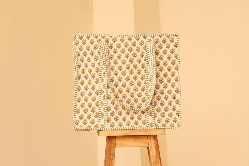 May include: A square tote bag with a floral pattern in yellow and white, featuring a repeating design of small flowers. The bag has green accents and handles, and is sitting on a small wooden stool.