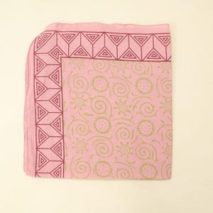 May include: A square pink fabric cloth featuring a dark pink geometric border pattern of triangles. The central area displays a repeating light green print of stylised suns and swirling lines across the light pink background surface.
