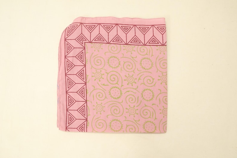 May include: A square pink fabric cloth featuring a dark pink geometric border pattern of triangles. The central area displays a repeating light green print of stylised suns and swirling lines across the light pink background surface.