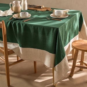 Op de afbeelding: Een groene en beige tafelkleed bedekt een tafel die gedekt is voor een maaltijd. Het tafelkleed is diepgroen met een brede beige rand. Borden, bestek en serveeronderdelen zijn op de tafel geplaatst. Een houten stoel en kruk zijn zichtbaar.