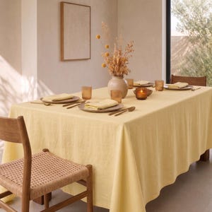 May include: A rectangular dining table covered with a solid pale yellow tablecloth, set with plates, folded napkins, amber-colored glassware, and gold-toned cutlery. A ceramic vase filled with dried flowers sits in the center of the table.
