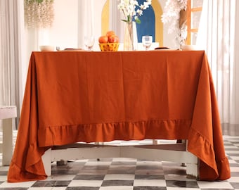 Puede incluir: Un mantel de color óxido con un borde con volantes cubre una mesa de madera blanca. La mesa está puesta con un cuenco, cubiertos, vasos y una cesta de naranjas. Un jarrón con flores blancas y una ventana están en el fondo.