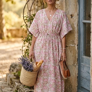 Op de afbeelding: Een vrouw draagt een roze jurk met bloemenprint, een V-hals en een taille met trekkoord. Ze draagt een gevlochten mand met lavendel en een baguette, en bruine leren schoenen. De jurk heeft korte mouwen en een vloeiend silhouet.