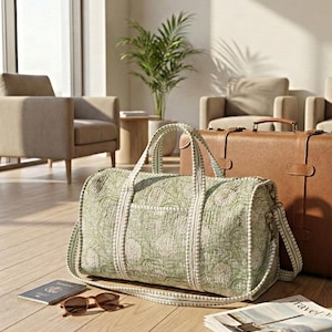 Può includere: Una borsa da viaggio verde con motivo floreale bianco, con tracolla e manici abbinati. Una valigia in pelle marrone è dietro la borsa. Occhiali da sole, passaporto e giornale sono sul pavimento in legno.