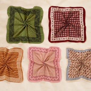 May include: Five square pocket squares arranged in a grid. Top left is olive green with a black border. Top right is deep red with a white floral pattern. Bottom left is brown with a geometric border. Bottom centre is pink with a dark pink border. Bottom right is cream with a brown and blue geometric pattern.