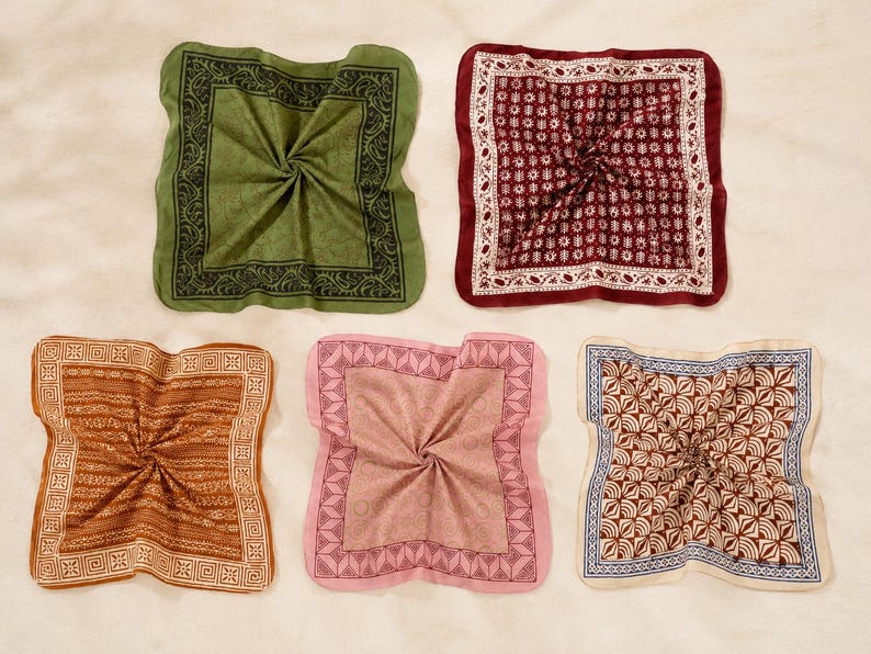 May include: Five square pocket squares arranged in a grid. Top left is olive green with a black border. Top right is deep red with a white floral pattern. Bottom left is brown with a geometric border. Bottom centre is pink with a dark pink border. Bottom right is cream with a brown and blue geometric pattern.
