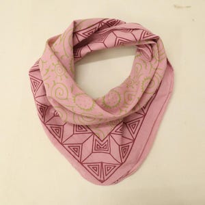 May include: A light pink bandana featuring a dark red geometric border pattern of triangles and lines, complemented by a central section decorated with olive green line art illustrations of suns, spirals, and circular shapes on the fabric.