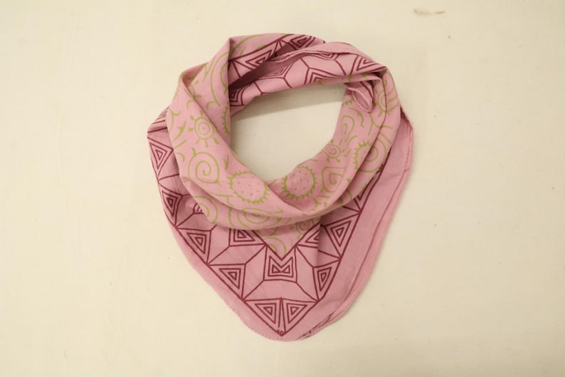 May include: A light pink bandana featuring a dark red geometric border pattern of triangles and lines, complemented by a central section decorated with olive green line art illustrations of suns, spirals, and circular shapes on the fabric.