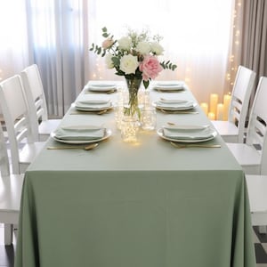 May include: A dining table set for a special occasion. The table is covered with a sage green tablecloth and set with plates, silverware, and napkins. A vase of flowers sits in the center, and white chairs surround the table.