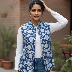 Puede incluir: Un chaleco azul sin mangas con un estampado floral blanco. El chaleco se lleva sobre una camisa blanca de manga larga y vaqueros azules. El chaleco tiene cuello mandarín y cierre de botones.