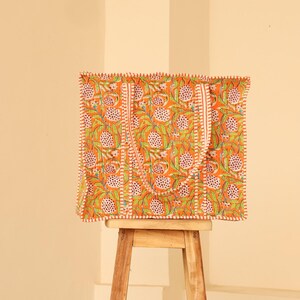 May include: Orange tote bag with a floral pattern in green and white. The bag has a rectangular shape with a matching handle and a white stitched border. The bag is sitting on a small wooden stool.