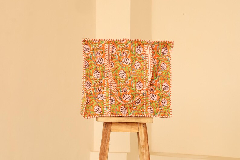 May include: Orange tote bag with a floral pattern in green and white. The bag has a rectangular shape with a matching handle and a white stitched border. The bag is sitting on a small wooden stool.