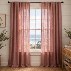 May include: A pair of floor-length, semi-sheer curtains in a dusty rose colour, featuring a textured, linen-like weave. The curtains are hung on a simple black rod, framing a window that looks out onto a coastal view with the ocean and trees.