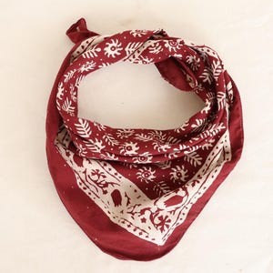 May include: A deep red bandana featuring a repeating white pattern of stylised suns and leafy branches. The fabric is folded into a triangular shape with a wide white border along the bottom edge, showcasing a detailed botanical print design.