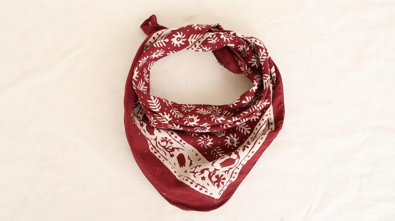 May include: A deep red bandana featuring a repeating white pattern of stylised suns and leafy branches. The fabric is folded into a triangular shape with a wide white border along the bottom edge, showcasing a detailed botanical print design.
