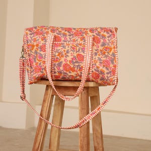 May include: A floral print tote bag with a shoulder strap, resting on a wooden stool. The bag features a repeating pattern of pink, orange, and gray flowers on a peach background. The straps are white with a red trim.