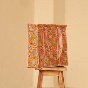May include: An orange tote bag with a floral pattern in green and white. The bag has a matching strap and is sitting on a small wooden stool. The bag's edges are trimmed with a white and red border.