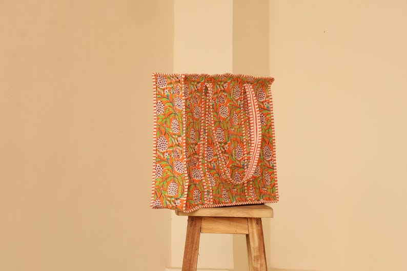 May include: An orange tote bag with a floral pattern in green and white. The bag has a matching strap and is sitting on a small wooden stool. The bag's edges are trimmed with a white and red border.