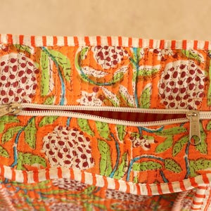 May include: Close-up of a quilted fabric with an orange background and a floral pattern in white, green, and maroon. The fabric has a white zipper and a striped border in orange and white.