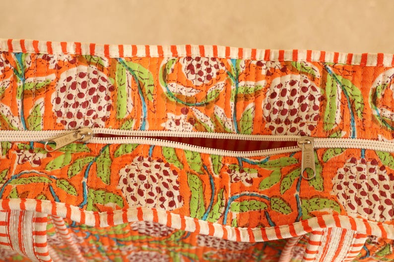 May include: Close-up of a quilted fabric with an orange background and a floral pattern in white, green, and maroon. The fabric has a white zipper and a striped border in orange and white.
