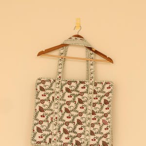 May include: Cream-colored tote bag with a floral pattern in red and green, hanging on a wooden hanger. The bag features a repeating design of stylized flowers and vines, with matching handles.