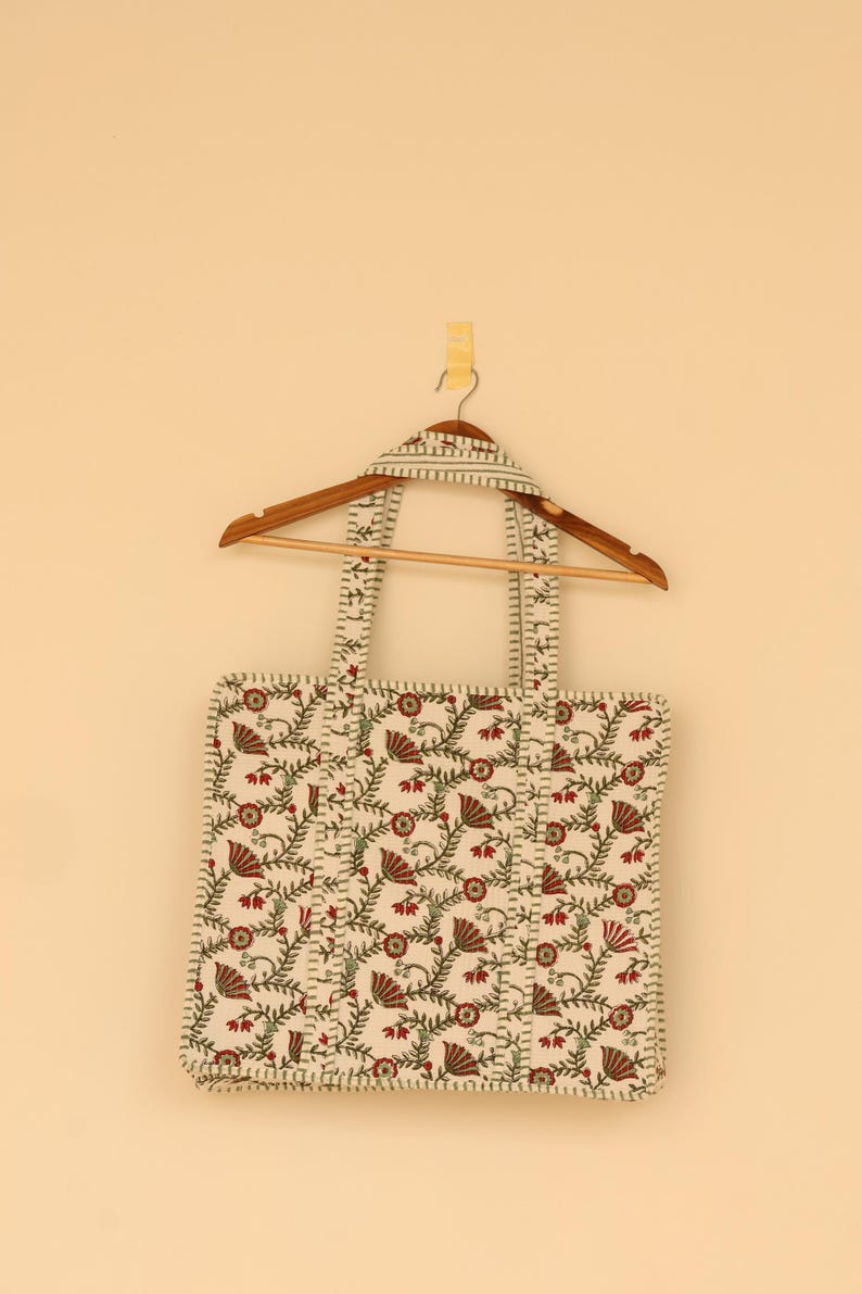 May include: Cream-colored tote bag with a floral pattern in red and green, hanging on a wooden hanger. The bag features a repeating design of stylized flowers and vines, with matching handles.