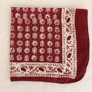 May include: A square fabric bandana in deep burgundy red featuring a repeating white block-printed pattern of stylised floral sprigs and sun-like motifs, framed by a decorative border of white tulips and vines on a solid burgundy red edge.