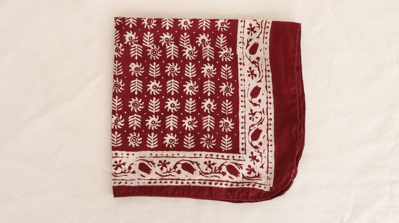 May include: A square fabric bandana in deep burgundy red featuring a repeating white block-printed pattern of stylised floral sprigs and sun-like motifs, framed by a decorative border of white tulips and vines on a solid burgundy red edge.