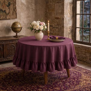 Op de afbeelding: Een ronde tafel met een dieppaarse tafelkleed met ruches. De tafel is gedekt met borden, servetten en een brandende kaars. Een vaas met bloemen staat op de tafel. De tafel staat in een kamer met een tapijt en een raam.