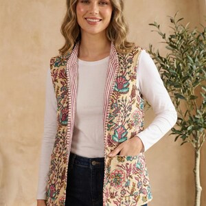Peut inclure: Un gilet sans manches crème avec un motif floral rose et turquoise. Le gilet a une doublure rayée rose et deux poches. Le modèle porte un haut blanc à manches longues et un jean bleu foncé.