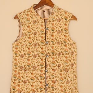 Puede incluir: Un chaleco sin mangas con un estampado floral en tonos amarillos, rojos y verdes. El chaleco tiene cuello mandar&iacute;n y forro a rayas marr&oacute;n y blanco. Se abrocha con botones y cuelga de una percha de madera.