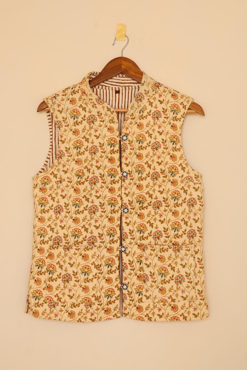 Puede incluir: Un chaleco sin mangas con un estampado floral en tonos amarillos, rojos y verdes. El chaleco tiene cuello mandar&iacute;n y forro a rayas marr&oacute;n y blanco. Se abrocha con botones y cuelga de una percha de madera.