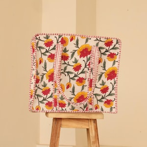 May include: A square tote bag with a floral pattern of yellow and red flowers on a white background. The bag has red stitching around the edges and handles. The bag is sitting on a small wooden stool.