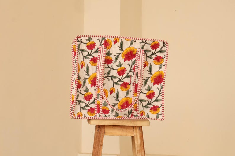 May include: A square tote bag with a floral pattern of yellow and red flowers on a white background. The bag has red stitching around the edges and handles. The bag is sitting on a small wooden stool.