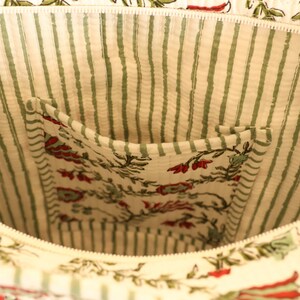 May include: Close-up of the interior of a cream-colored tote bag. The lining features green and white vertical stripes. A pocket is visible, showcasing a floral pattern with red and green accents. The bag has a zipper closure.