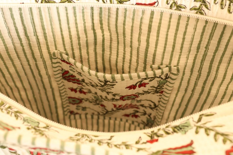 May include: Close-up of the interior of a cream-colored tote bag. The lining features green and white vertical stripes. A pocket is visible, showcasing a floral pattern with red and green accents. The bag has a zipper closure.