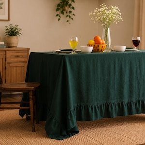Könnte beinhalten: Eine dunkelgrüne Tischdecke aus Leinen mit Rüschensaum bedeckt einen Holztisch. Der Tisch ist mit Obst, Gläsern und weißen Schalen gedeckt. Ein Holzstuhl steht neben dem Tisch, und ein gewebter Teppich liegt auf dem Boden. Eine Lampe und ein Schrank befinden sich im Hintergrund.