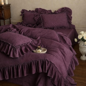 May include: A deep purple bedding set featuring a duvet cover and two matching pillowcases, all adorned with wide, gathered ruffles along the edges. The fabric has a visible, textured weave, creating a soft, layered appearance across the bed.