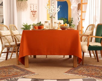 Puede incluir: Un comedor con un mantel naranja sobre una mesa de madera, rodeado de sillas de ratán con cojines crema. La escena incluye plantas, flores y pequeñas alfombras, creando un ambiente cálido y acogedor.