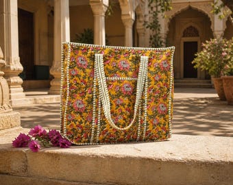 Op de afbeelding: Een grote, rechthoekige draagtas met een bloemenpatroon in geel, roze en rood. De tas heeft wit en groen gestreepte handvatten en randen. De tas staat op een stenen oppervlak met paarse bloemen.