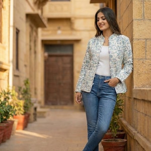 May include: A woman wearing a light blue and white patterned jacket, a white top, and blue jeans. The jacket has a stand-up collar. She is wearing sandals and is leaning against a sandstone wall in an outdoor setting.