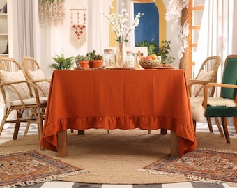 Puede incluir: Un comedor con una mesa cubierta con un mantel naranja con borde con volantes. Sillas de mimbre con cojines rodean la mesa. Alfombras decorativas y plantas realzan la habitación. Un jarrón con flores blancas está sobre la mesa.